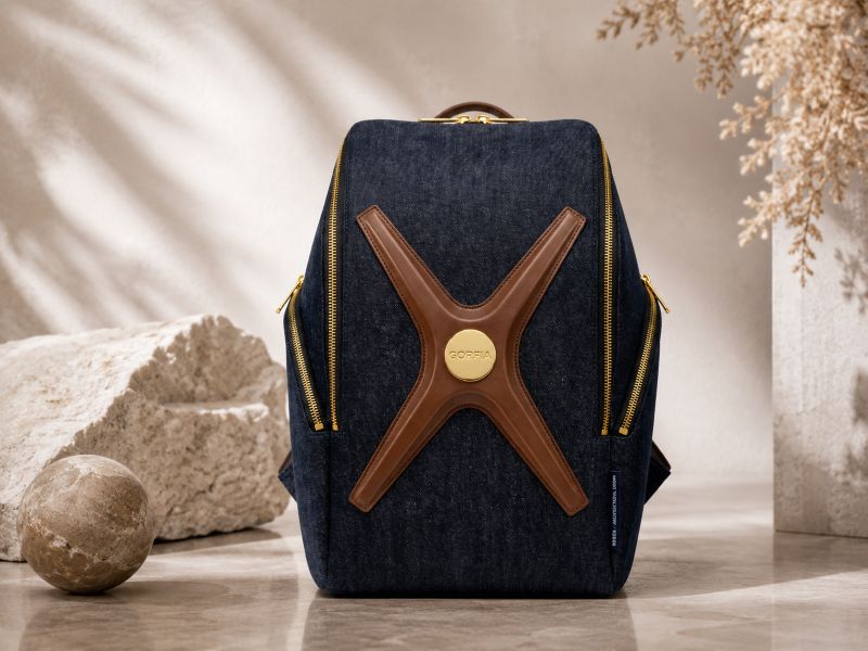 dark-indigo-denim-backpack-with-cross-shaped-brown-leather-strap-and-round-gold-metal-hardware-displayed-on-minimalist-beige-studio-background-with-stones-and-dried-fern-decorations